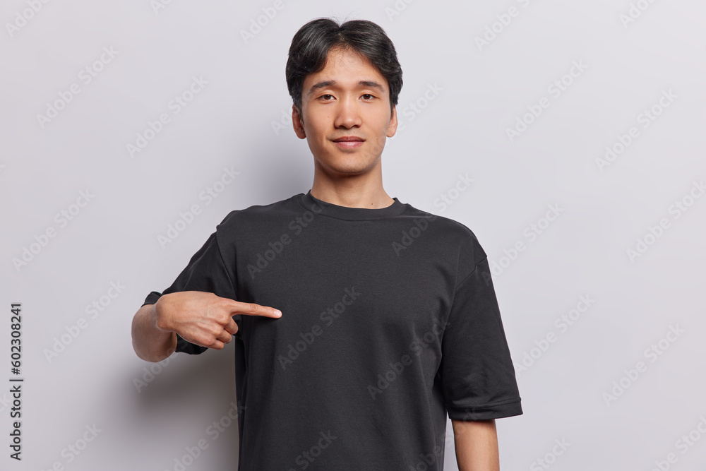 Wall mural Indoor shot of handsome brunet Asian man points with index finger at blank black tshirt empty space for your advertising text isolated over white background. This is me. People and clothing concept - Wall murals