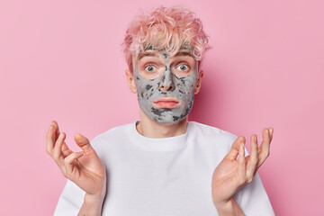 Clueless adult man spreads palms looks surprisingly at camera applies homemade clay mask on face to unclog pores stands hesitant indoor dressed in white t shirt isolated over pink background