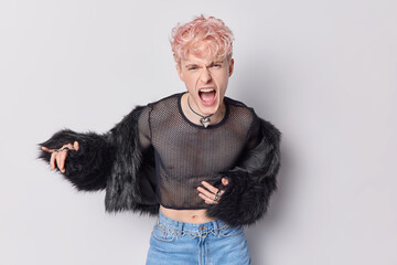 Indoor shot of annoyed pink haired man gay exclaims loudly wears net jumper fur fahionable coat and jeans feels annoyed isolated over white background. Non binary people and emotions concept