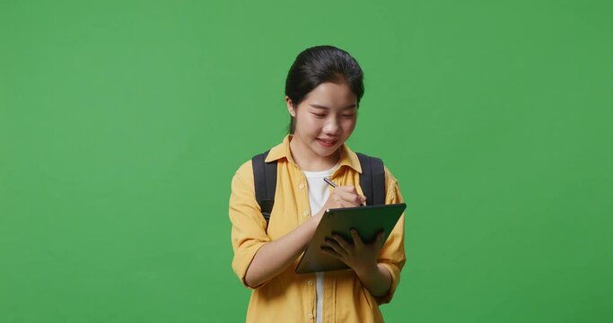 Close Up Of Asian Woman Student With A Backpack Smiling And Taking Note On The Tablet While Standing In The Green Screen Background Studio
