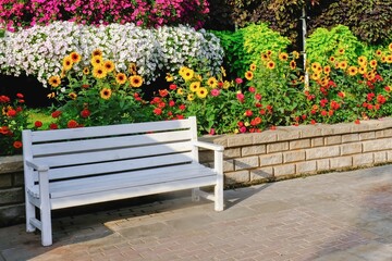 White wooden bench beside path in garden with blossom flowers and trees in background. Bench in public garden. Urban street furniture