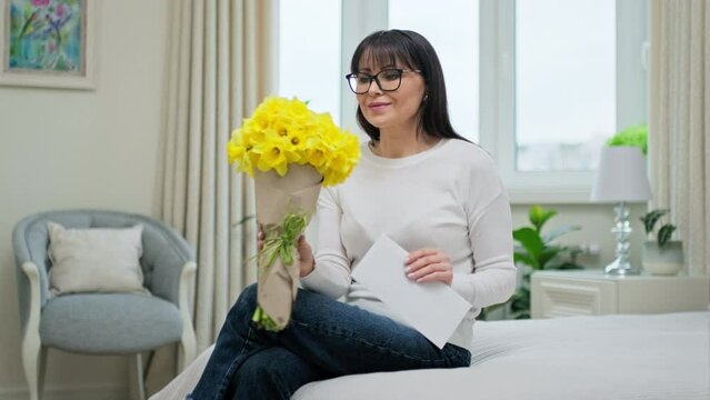 Happy middle aged female with bouquet of flowers, reading congratulations note, at home