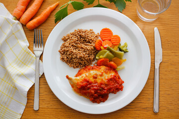Chicken breast steak with mozzarella and tomato salsa served with egg barley (tarhona) and boiled vegetables on a white plate.