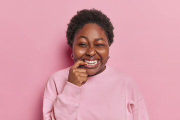 Studio shot of cheerful dark skinned woman bites finger winks eye and feels happy winks eye dressed...
