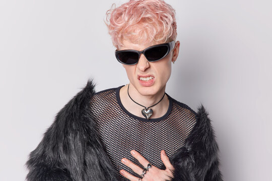 Handsome man with trnedy pink hairstyle clenches teeth has annoyed expression wears trendy sunglasses and black fur coat isolated over white background. Sweet guy portrait. Androgynous male.