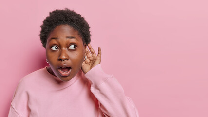 Curious dark skinned woman keeps hand near ear tries to overhear listens secrets eavesdropes interesting news keeps mouth opened wears casual jumper isolated over pink wall copy space for your text - obrazy, fototapety, plakaty