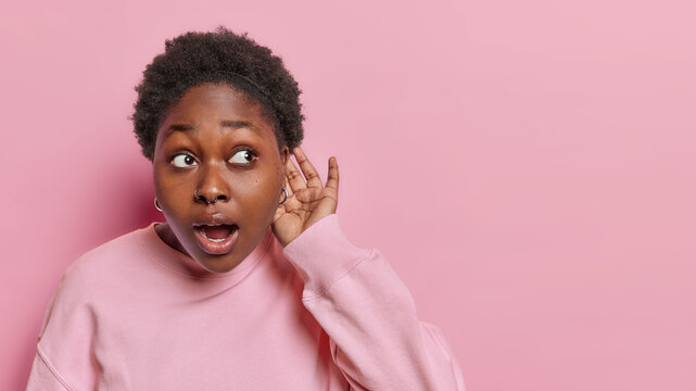 Curious dark skinned woman keeps hand near ear tries to overhear listens secrets eavesdropes interesting news keeps mouth opened wears casual jumper isolated over pink wall copy space for your text