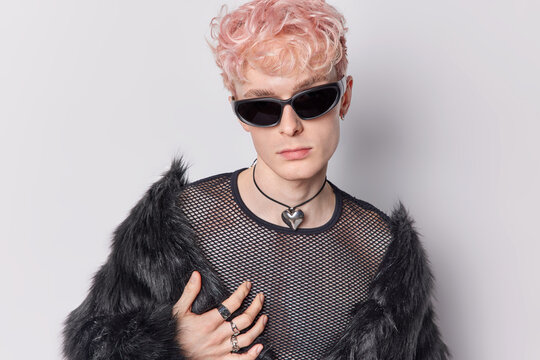Portrait of handsome androgynous man with pink curly hair wears sunglasses dressed in black fur coat and net jumper stands self confident indoor against white background. Transgender male model