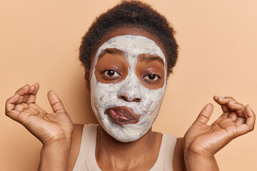 Hesitant short haired woman with dark skin applies white beauty mask on face to improve skin...