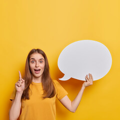 Impressed dark haired woman has idea or answer on question keeps index finger raised up holds blank speech bubble for your advertising content dressed in casual t shirt isolated over yellow background