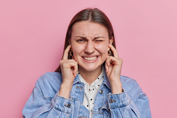 Irritated dark haired young pretty European woman plugs ears with fingers grimaces from loud noise avoids hearing something dressed in denim jacket isolated over pink background. I cant stand this - obrazy, fototapety, plakaty