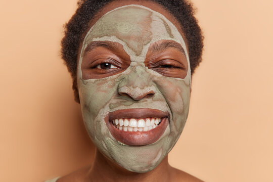 Close up image of Afro woman frowns face and clenches teeth applies nourishing clay mask with cleansing properties to regulate oil secretion and balance has funny expression isolated over brown wall