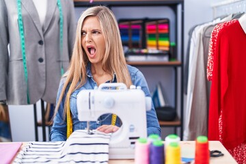 Naklejka na ściany i meble Blonde woman dressmaker designer using sew machine angry and mad screaming frustrated and furious, shouting with anger. rage and aggressive concept.