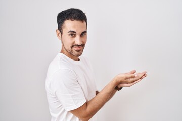 Handsome hispanic man standing over white background pointing aside with hands open palms showing copy space, presenting advertisement smiling excited happy