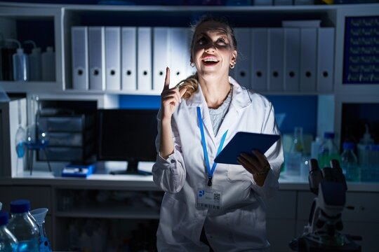 Beautiful blonde woman working at scientist laboratory late at night amazed and surprised looking up and pointing with fingers and raised arms.