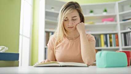 Young blonde woman student reading book sitting on table at library university