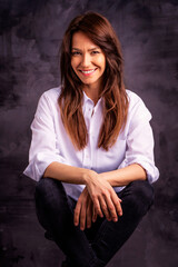 Studio portrait of a brunette haired attractive woman wearing white shirt and black jeans while posing at isolated dark background