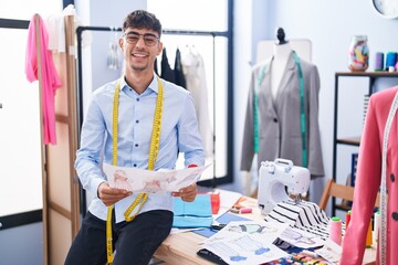 Young hispanic man tailor smiling confident looking clothing design at tailor shop