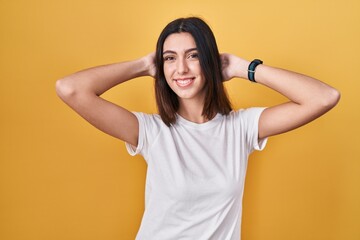 Young beautiful woman standing over yellow background relaxing and stretching, arms and hands behind head and neck smiling happy