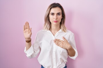 Young beautiful woman standing over pink background swearing with hand on chest and open palm, making a loyalty promise oath