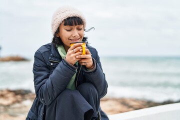 Young beautiful hispanic woman smiling confident drinking coffee at seaside