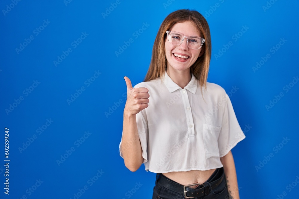 Sticker Beautiful woman standing over blue background doing happy thumbs up gesture with hand. approving expression looking at the camera showing success.