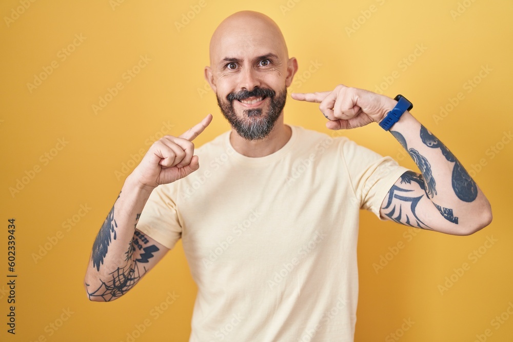 Wall mural Hispanic man with tattoos standing over yellow background smiling cheerful showing and pointing with fingers teeth and mouth. dental health concept.