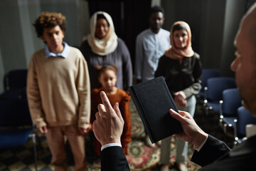 Priest holding book with prayers and reading sermon for people while they standing in church
