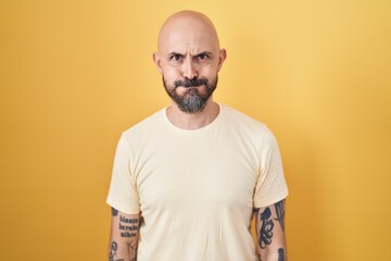 Hispanic man with tattoos standing over yellow background puffing cheeks with funny face. mouth inflated with air, crazy expression.