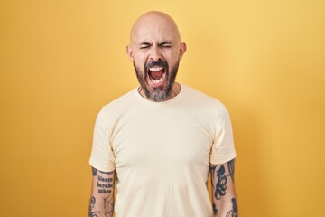 Hispanic man with tattoos standing over yellow background angry and mad screaming frustrated and furious, shouting with anger. rage and aggressive concept.