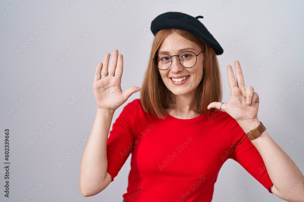 Wall mural young redhead woman standing wearing glasses and beret showing and pointing up with fingers number e