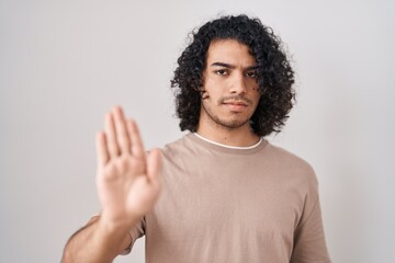 Hispanic man with curly hair standing over white background doing stop sing with palm of the hand. warning expression with negative and serious gesture on the face.