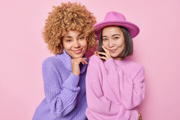 Portrait of two female friends stand closely to each other have natural gentle smile wear knitted jumpers hat isolated over pink wall meet together at weekend. Beautiful women in fashionable clothes