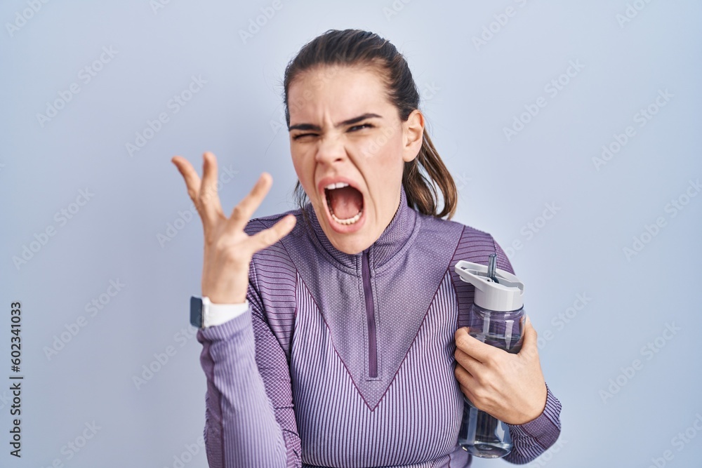 Poster beautiful woman wearing sportswear holding water bottle crazy and mad shouting and yelling with aggr