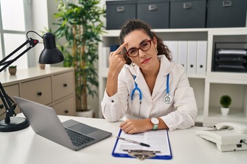 Young hispanic woman wearing doctor uniform and stethoscope pointing unhappy to pimple on forehead, ugly infection of blackhead. acne and skin problem