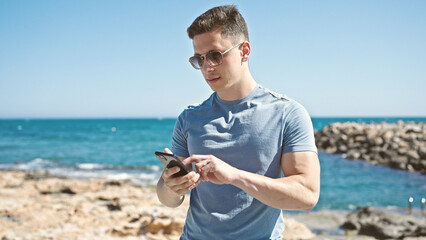 Young hispanic man tourist using smartphone with relaxed expression at seaside