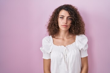 Fototapeta premium Hispanic woman with curly hair standing over pink background relaxed with serious expression on face. simple and natural looking at the camera.