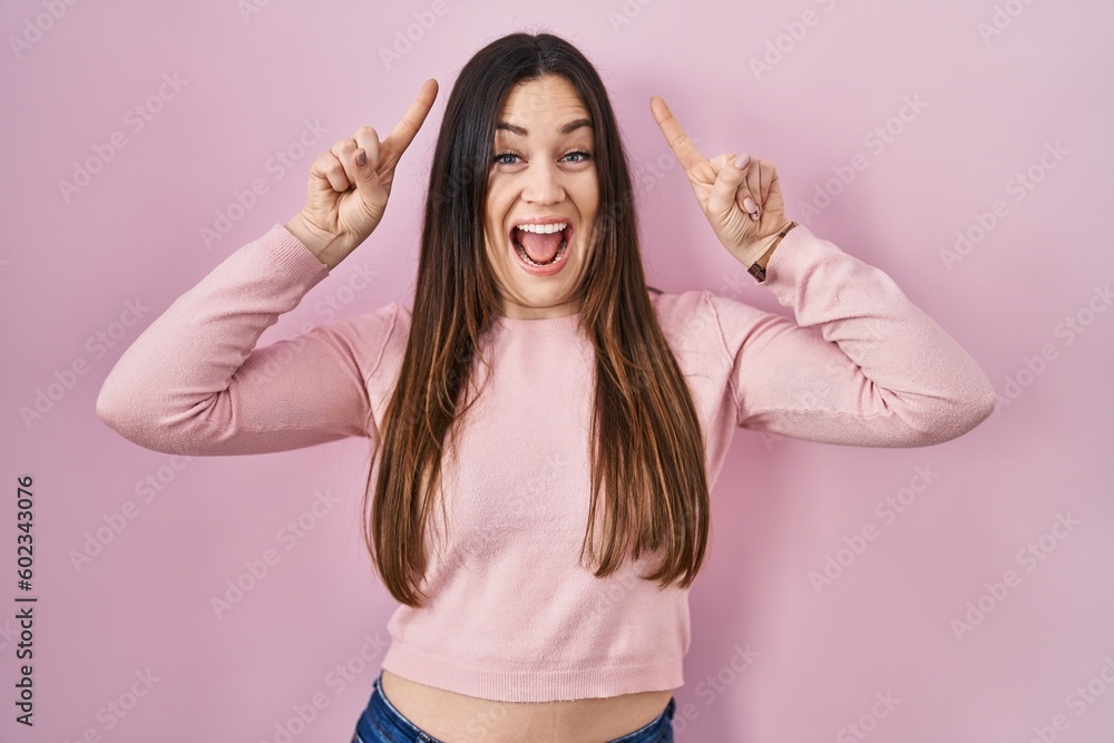 Sticker Young brunette woman standing over pink background smiling amazed and surprised and pointing up with fingers and raised arms.