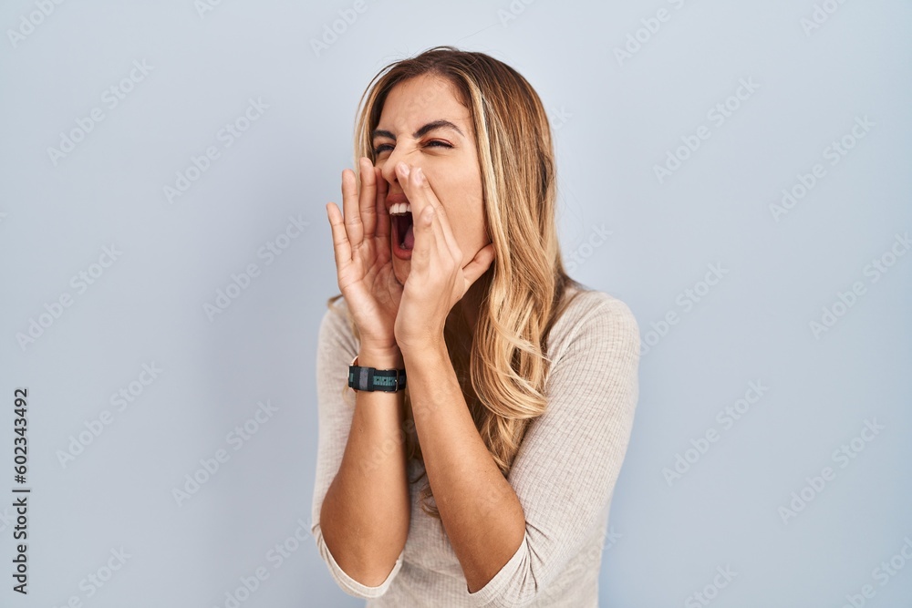 Wall mural young blonde woman standing over isolated background shouting angry out loud with hands over mouth