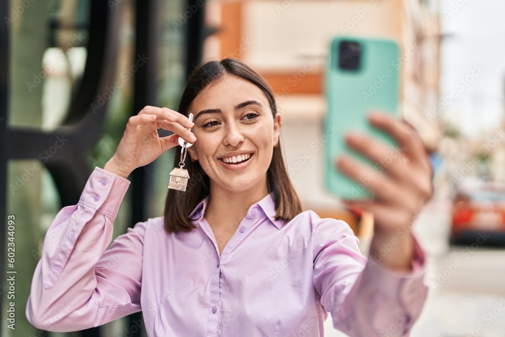 Wall mural Young hispanic woman make selfie by the smartphone holding key of new home at street