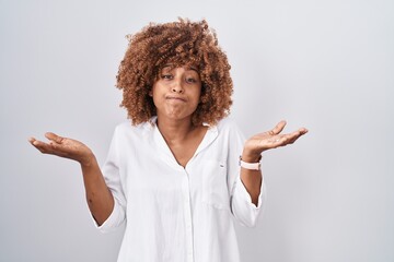 Young hispanic woman with curly hair standing over white background clueless and confused expression with arms and hands raised. doubt concept.