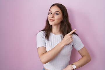 Young hispanic girl standing over pink background pointing with hand finger to the side showing...
