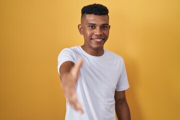 Young hispanic man standing over yellow background smiling friendly offering handshake as greeting and welcoming. successful business.