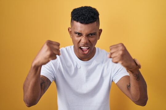 Young hispanic man standing over yellow background angry and mad raising fists frustrated and furious while shouting with anger. rage and aggressive concept.