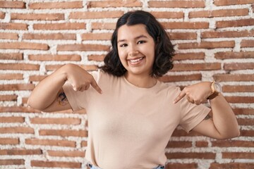 Young hispanic woman standing over bricks wall looking confident with smile on face, pointing...