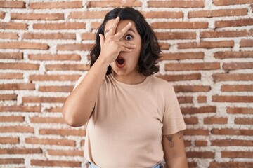 Fototapeta na wymiar Young hispanic woman standing over bricks wall peeking in shock covering face and eyes with hand, looking through fingers with embarrassed expression.