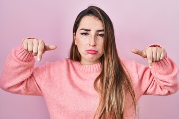 Young hispanic woman standing over pink background pointing down looking sad and upset, indicating...