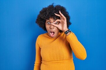 Fototapeta na wymiar Black woman with curly hair standing over blue background doing ok gesture shocked with surprised face, eye looking through fingers. unbelieving expression.