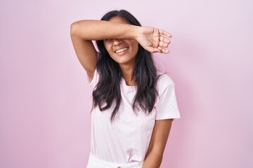 Young hispanic woman standing over pink background covering eyes with arm smiling cheerful and funny. blind concept.