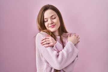 Young caucasian woman standing over pink background hugging oneself happy and positive, smiling...